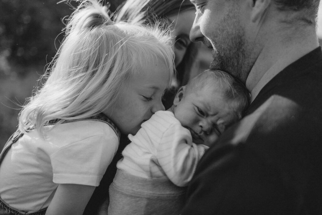 Familienfotografie Hamburg. Mädchen küsst liebevoll Neugeborenes, das auf dem Arm der Eltern liegt.