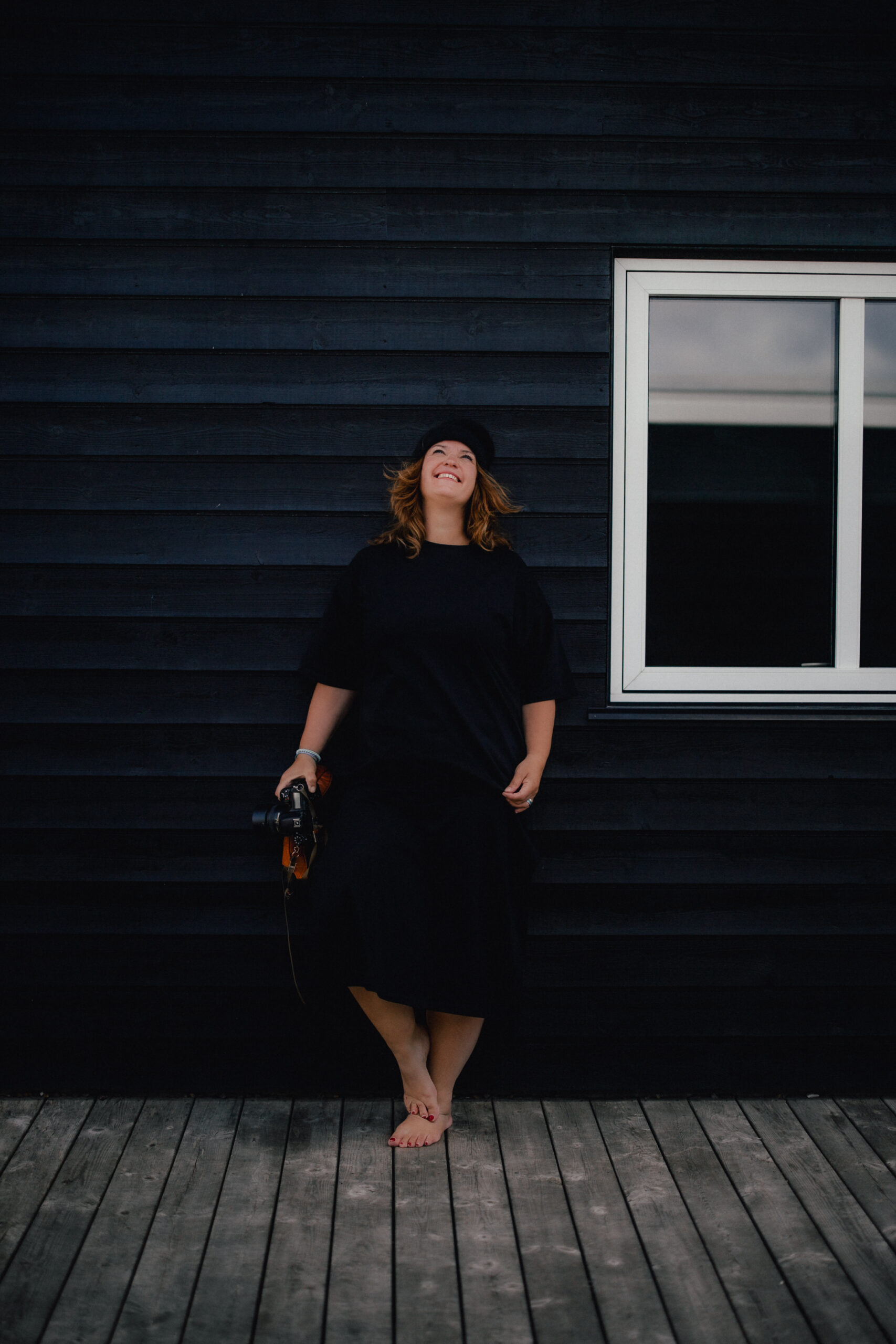 Angie Hagedorn, Hochzeitsfotografin, lehnt sich lächelnd an eine dunkelblaue Hausfassade und hält eine Kamera in der Hand.